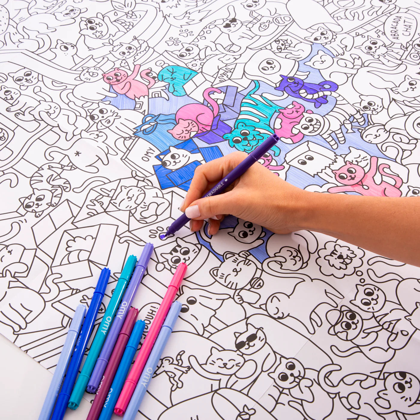 Coloring book with cat illustrations and a hand using a purple pen to color.