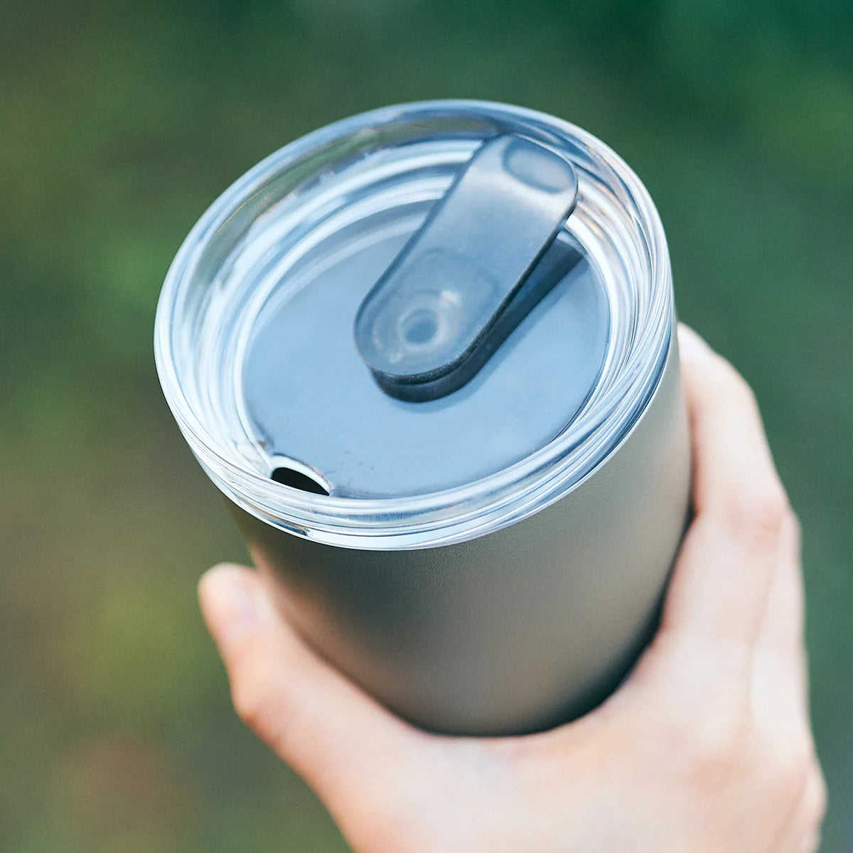 Hand holding a greeninsulated tumbler with a clear lid against a blurred green background