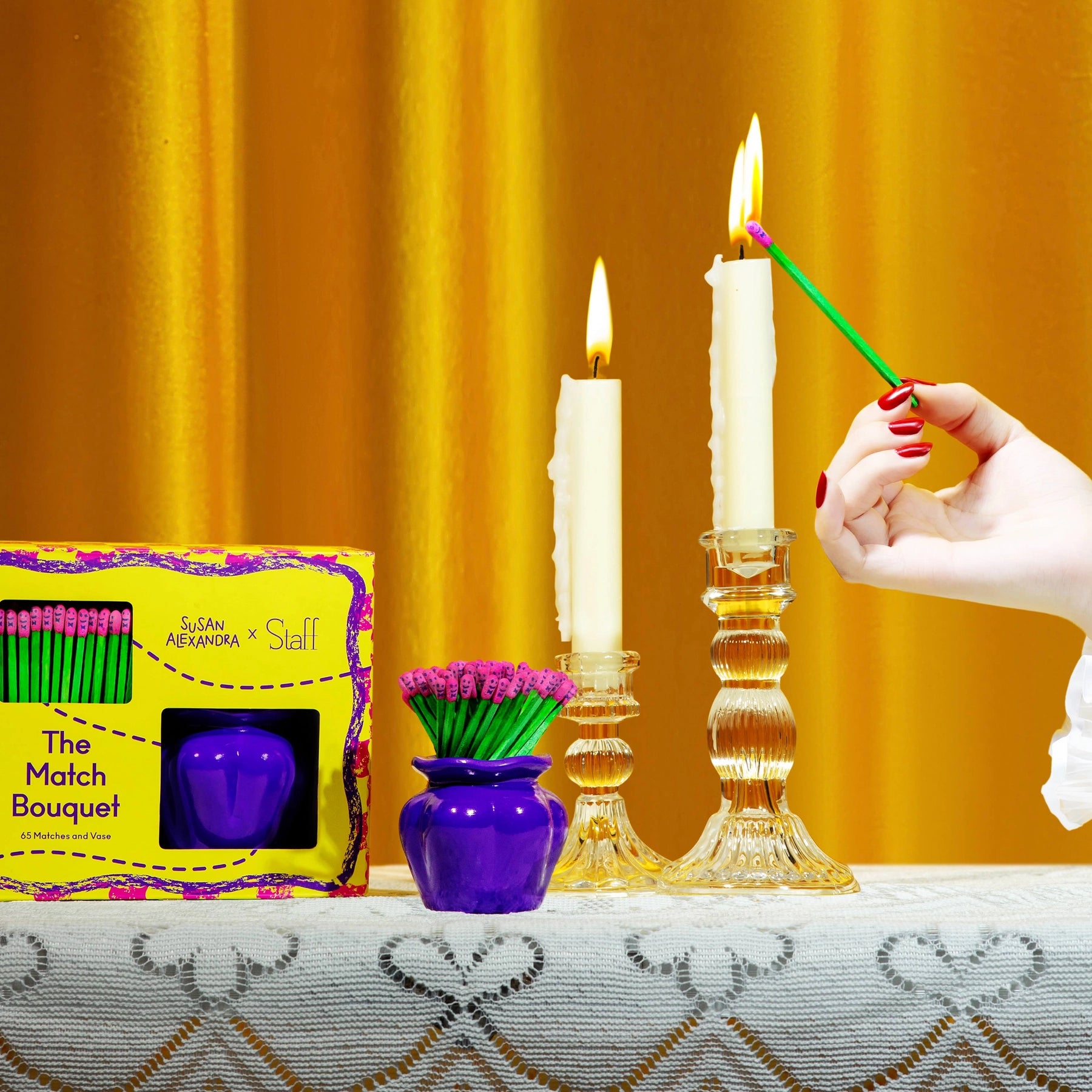 Candle being lit with a match, next to a box of matches and a vase with purple flowers on a yellow background.