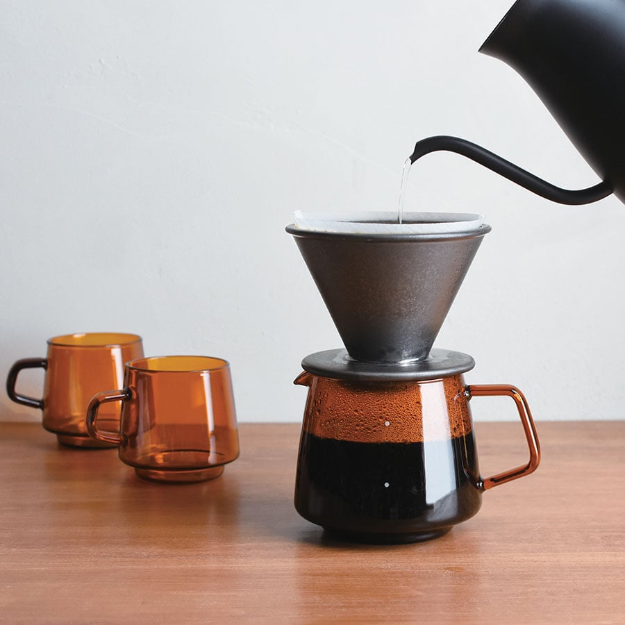 Coffee being poured from a kettle into a coffee filter on a wooden table with two orange cups.