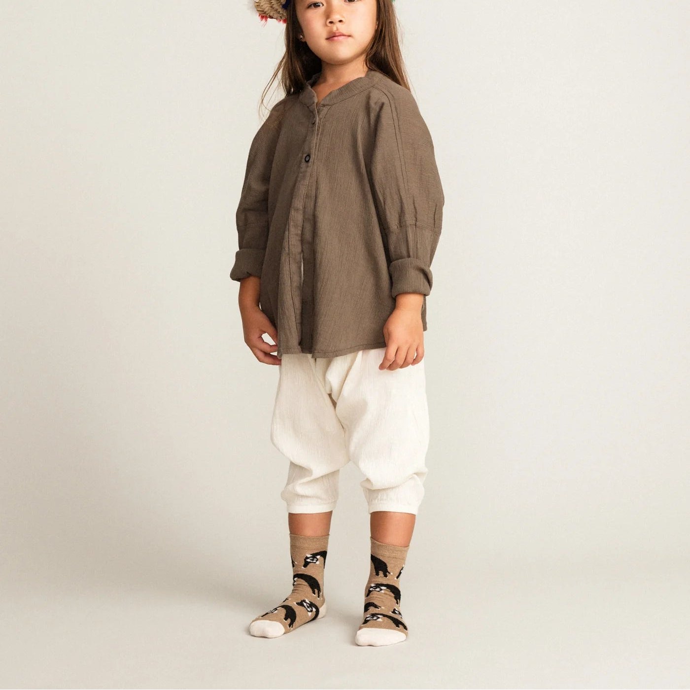 Child wearing a brown shirt, white pants, and a colorful hat on a light gray background