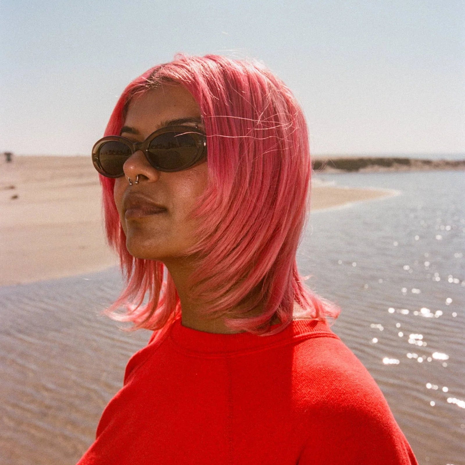Person with pink hair and sunglasses standing by a body of water