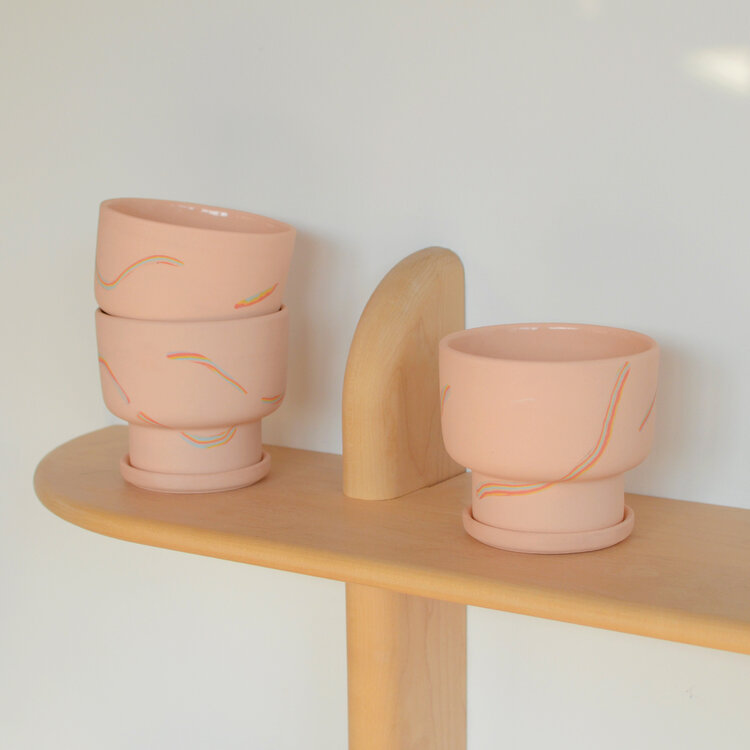 Set of pink ceramic cups with abstract designs on a wooden shelf against a white wall.