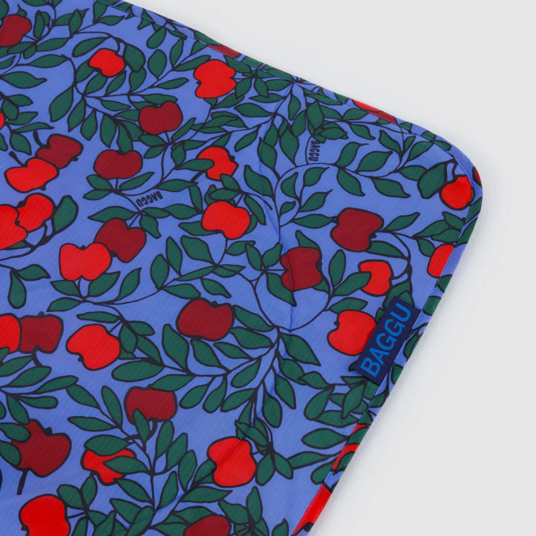 Corner of a blanket with red apple pattern on a blue background, featuring the brand 'Baggu'.