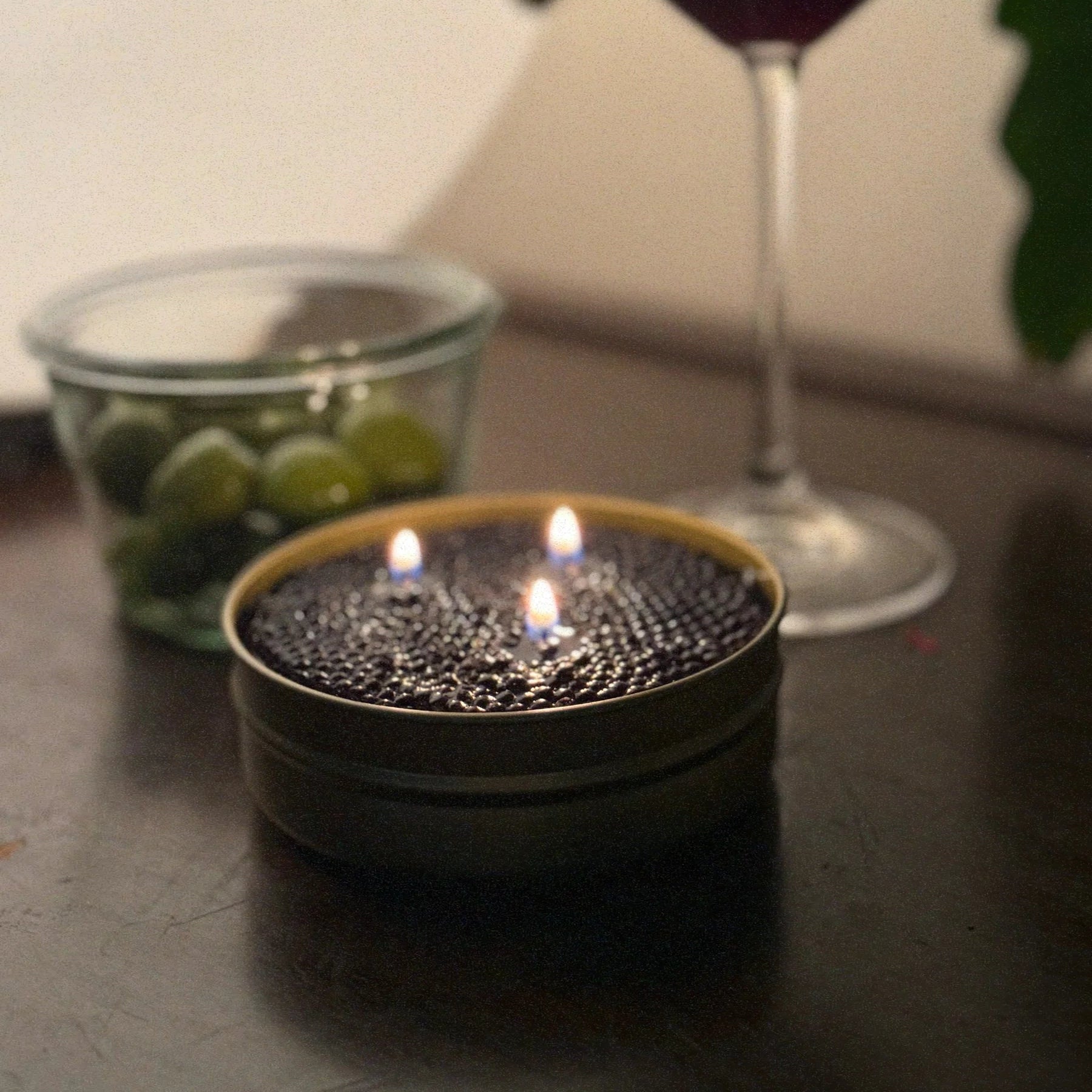 Candle in a tin with two lit wicks, glass of red wine, and bowl of green olives on a dark surface.
