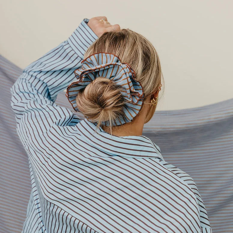A striped fabric scrunchie with blue, brown, and white horizontal stripes, featuring a white paper tag with the brand name 'kaxi' attached, set against a plain white background.