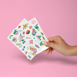 Hand holding two sheets of colorful unicorn-themed tattoos against a pink background