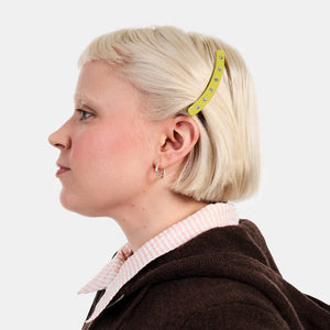 Person with a yellow hair clip wearing a brown jacket and pink shirt on a white background