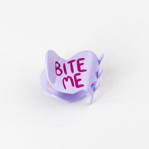 Purple heart-shaped hair claw with 'BITE ME' text on a white background