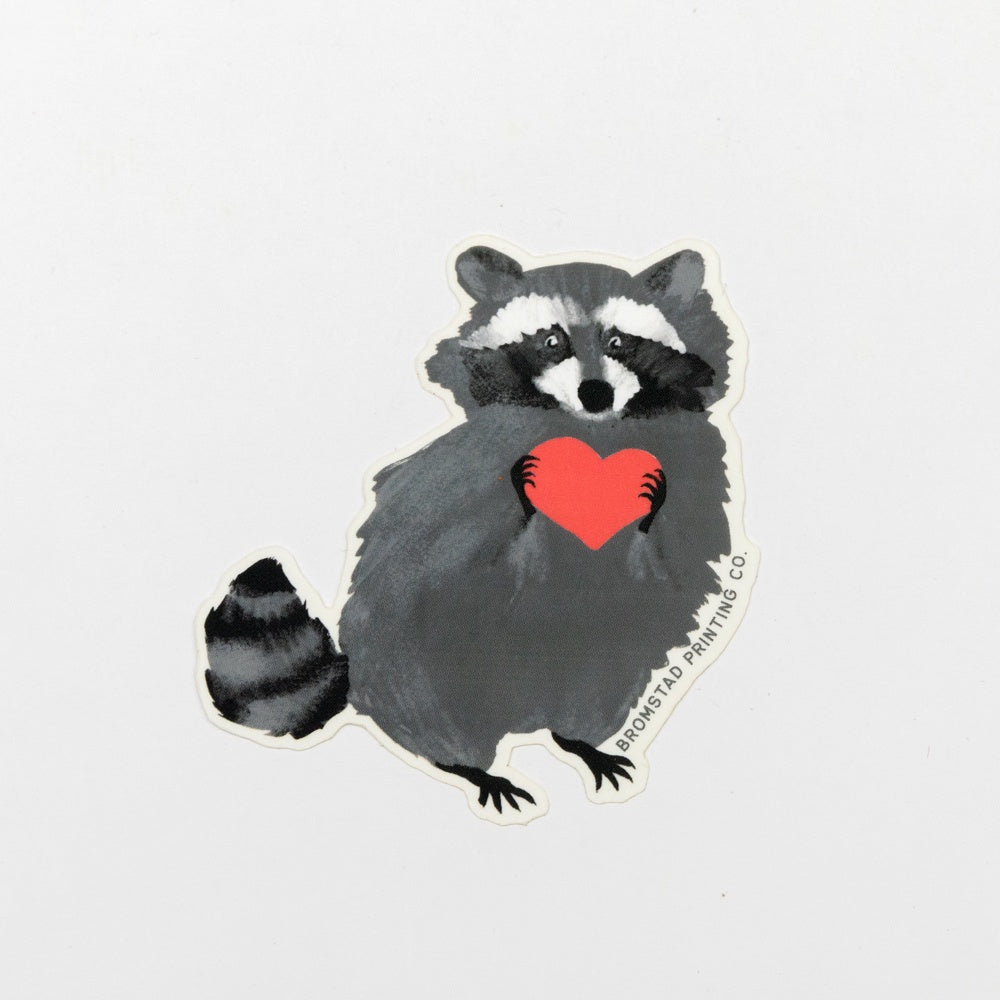 Sticker of a raccoon holding a red heart on a light gray background