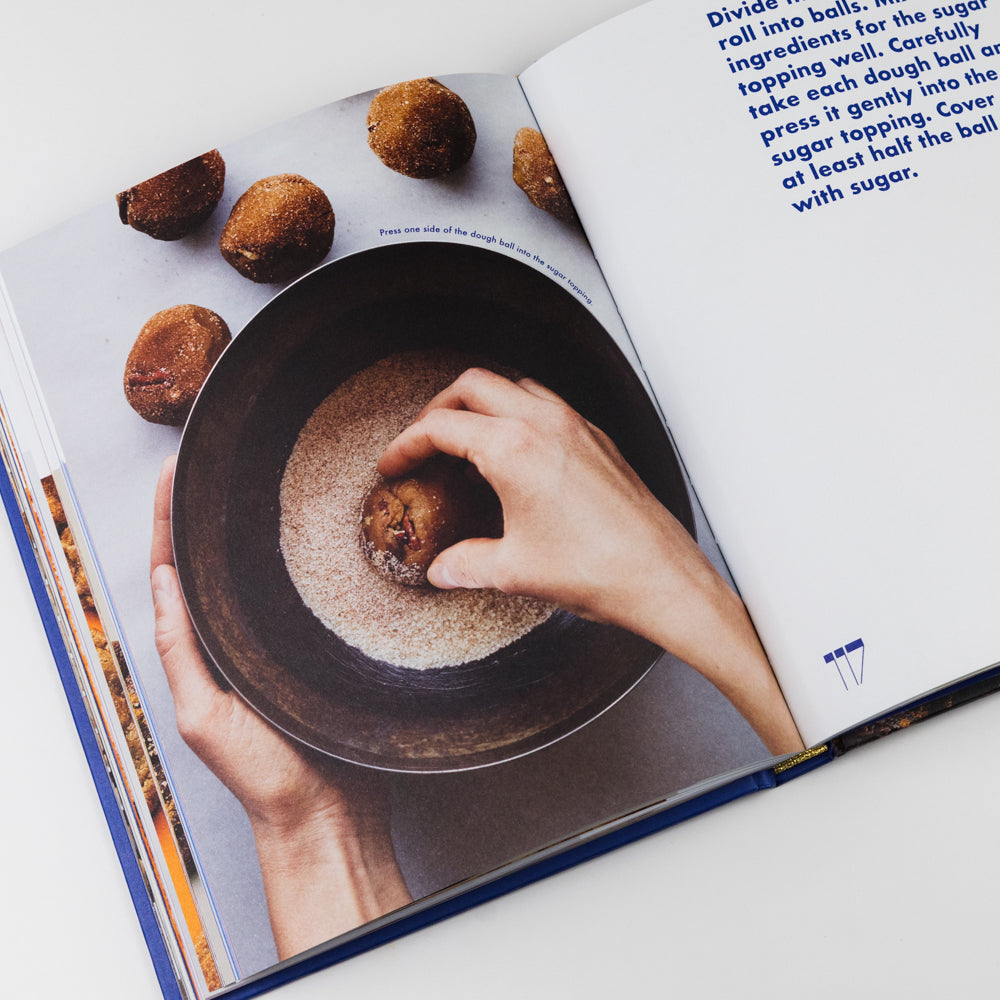 Open cookbook showing a hand rolling a dough ball in sugar over a dark surface.