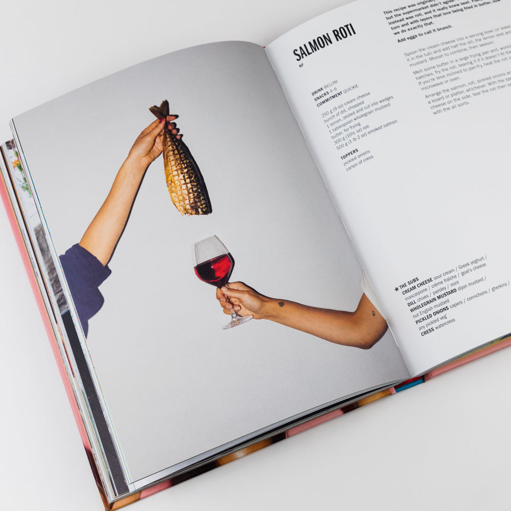 Open cookbook showing a page with two hands holding a glass of red wine and a headless fish, on a white background.