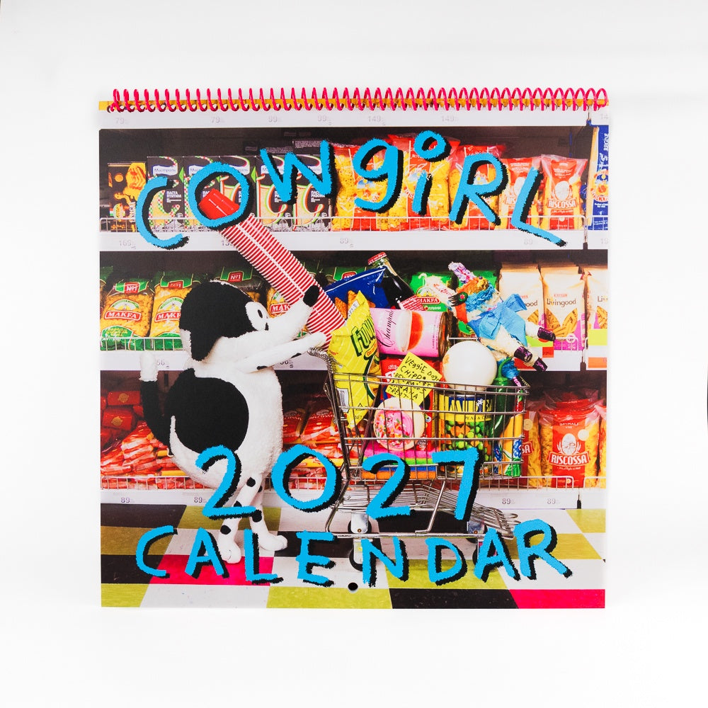 A spiral-bound 2027 wall calendar titled 'Cowgirl' featuring a colorful grocery store scene with a black-and-white dog pushing a shopping cart, photographed against a plain white background.