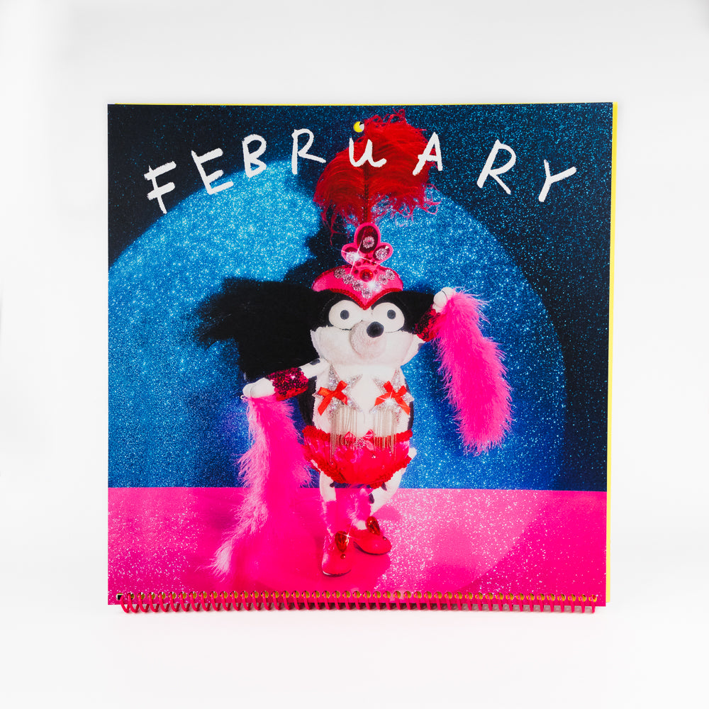 A spiral-bound 2027 wall calendar titled 'Cowgirl' featuring a colorful grocery store scene with a black-and-white dog pushing a shopping cart, photographed against a plain white background.
