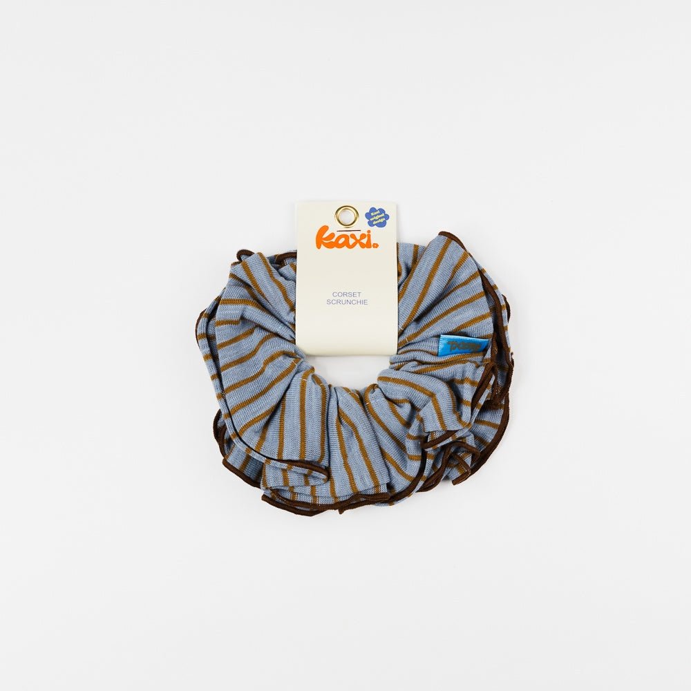 A striped fabric scrunchie with blue, brown, and white horizontal stripes, featuring a white paper tag with the brand name 'kaxi' attached, set against a plain white background.