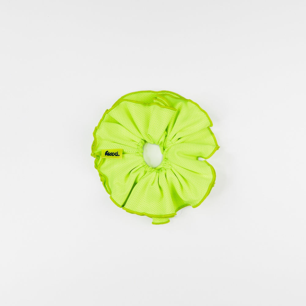 A bright lime green fabric scrunchie with a small black and yellow 'kaxi.' label, photographed flat on a clean white background.