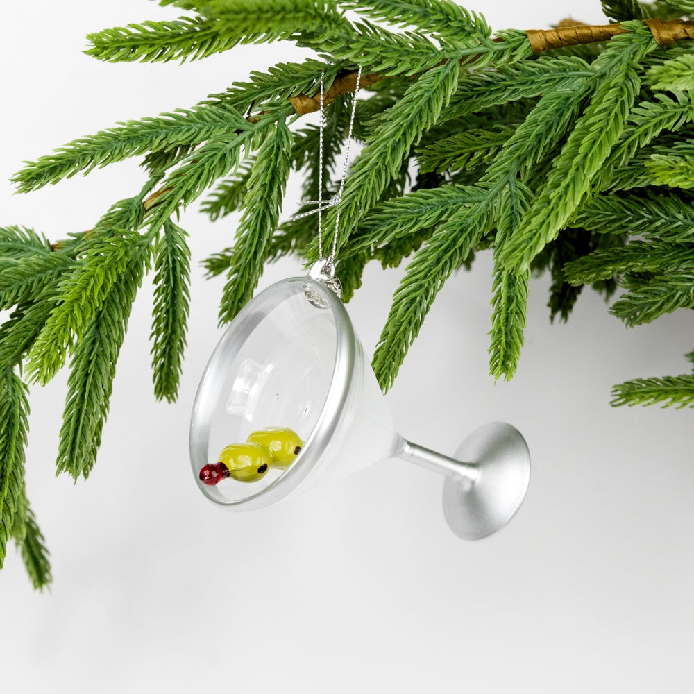 Decorative martini ornament with two olives on a toothpick hanging from a branch against a white background.