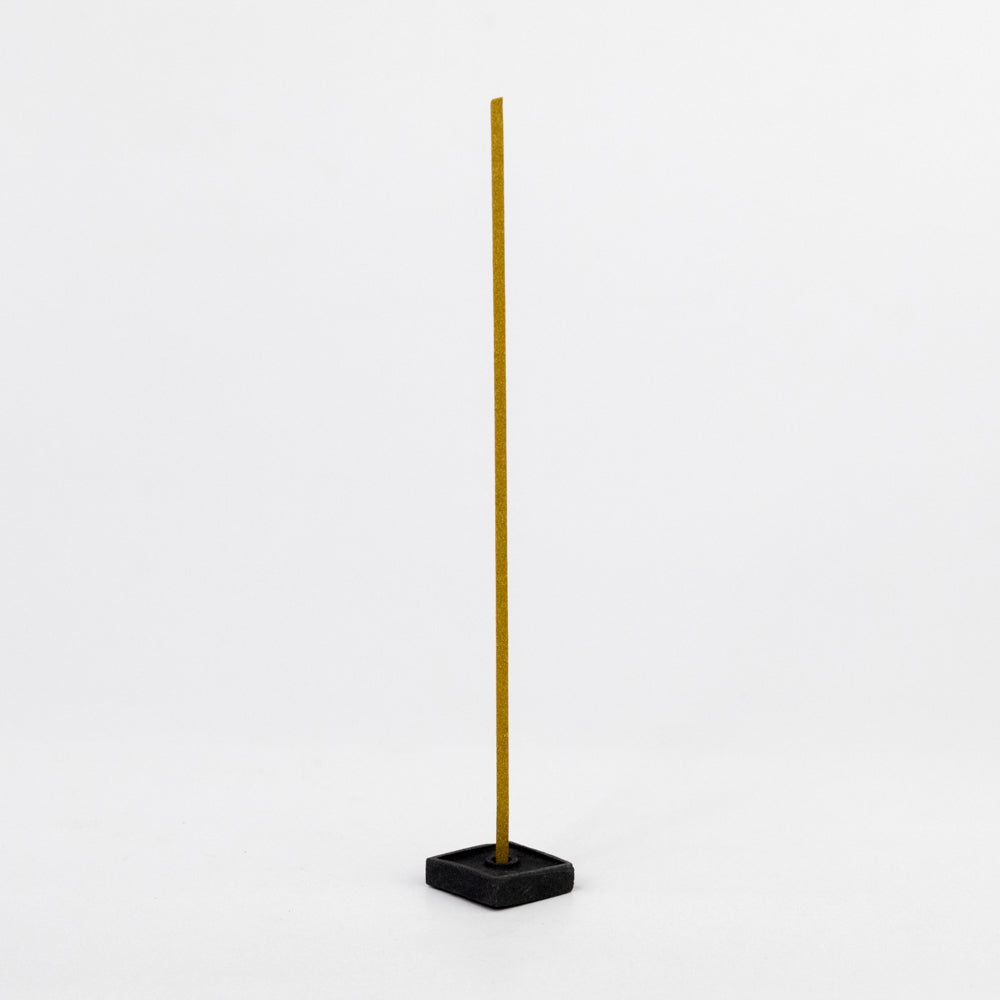 Tall, thin yellow incense stick with a black square holder on a white background