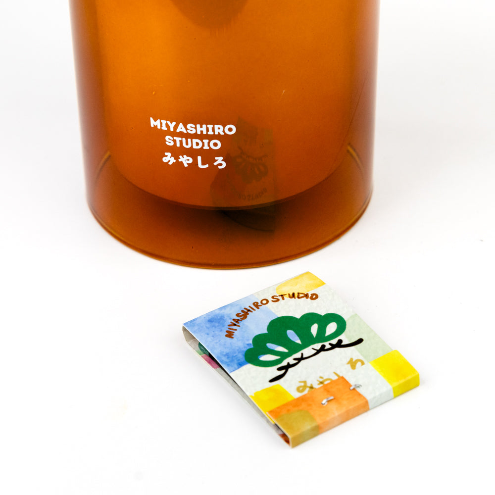 Orange glass with 'MIYASHIRO STUDIO' branding next to a colorful matchbook on a white background