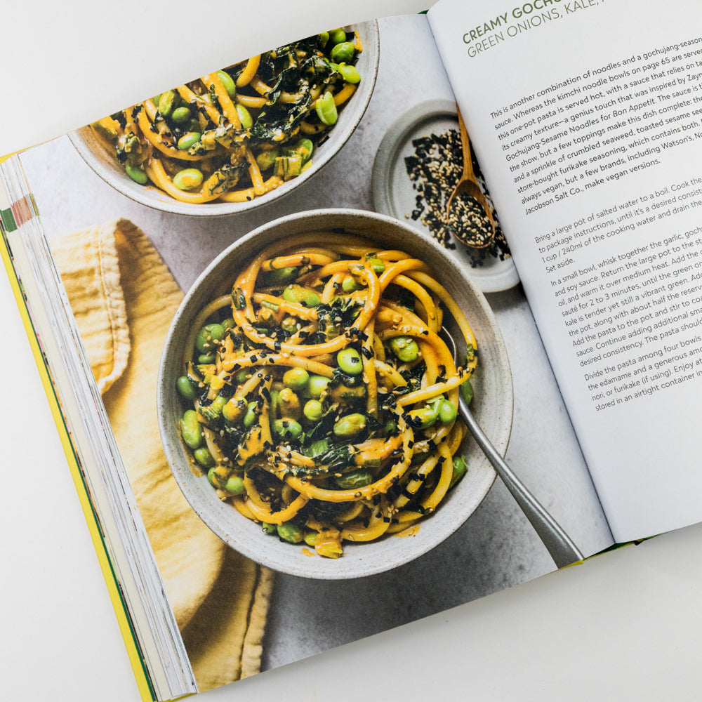 Open cookbook showing a recipe for creamy gochujang green onions and edamame with a bowl of the dish on a white surface.