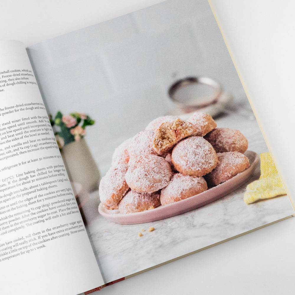 Open cookbook showing a plate of powdered sugar-covered pastries on a marble surface.