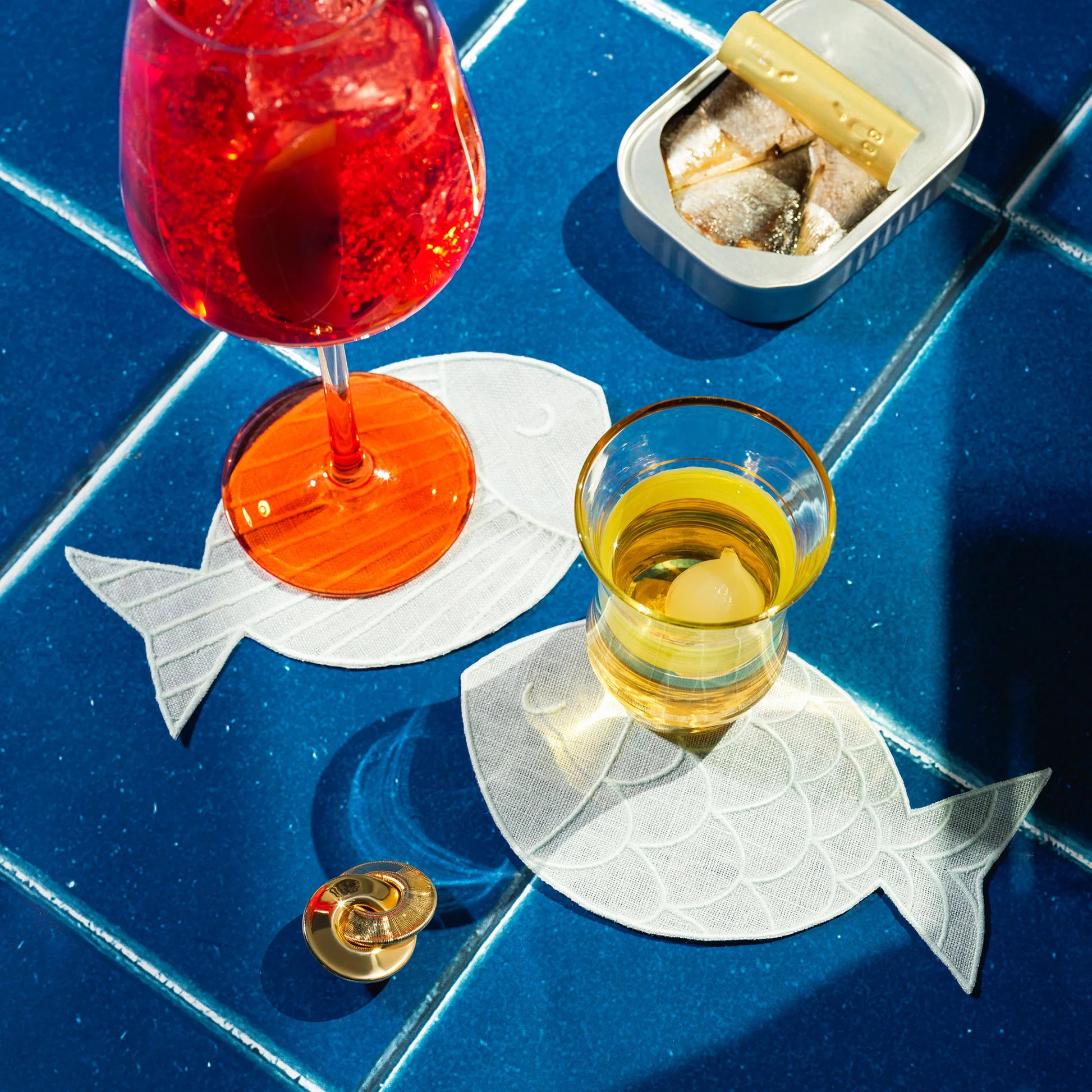 Two glasses of red and yellow drinks on fish-shaped coasters with a small container of snacks on a blue tiled surface.