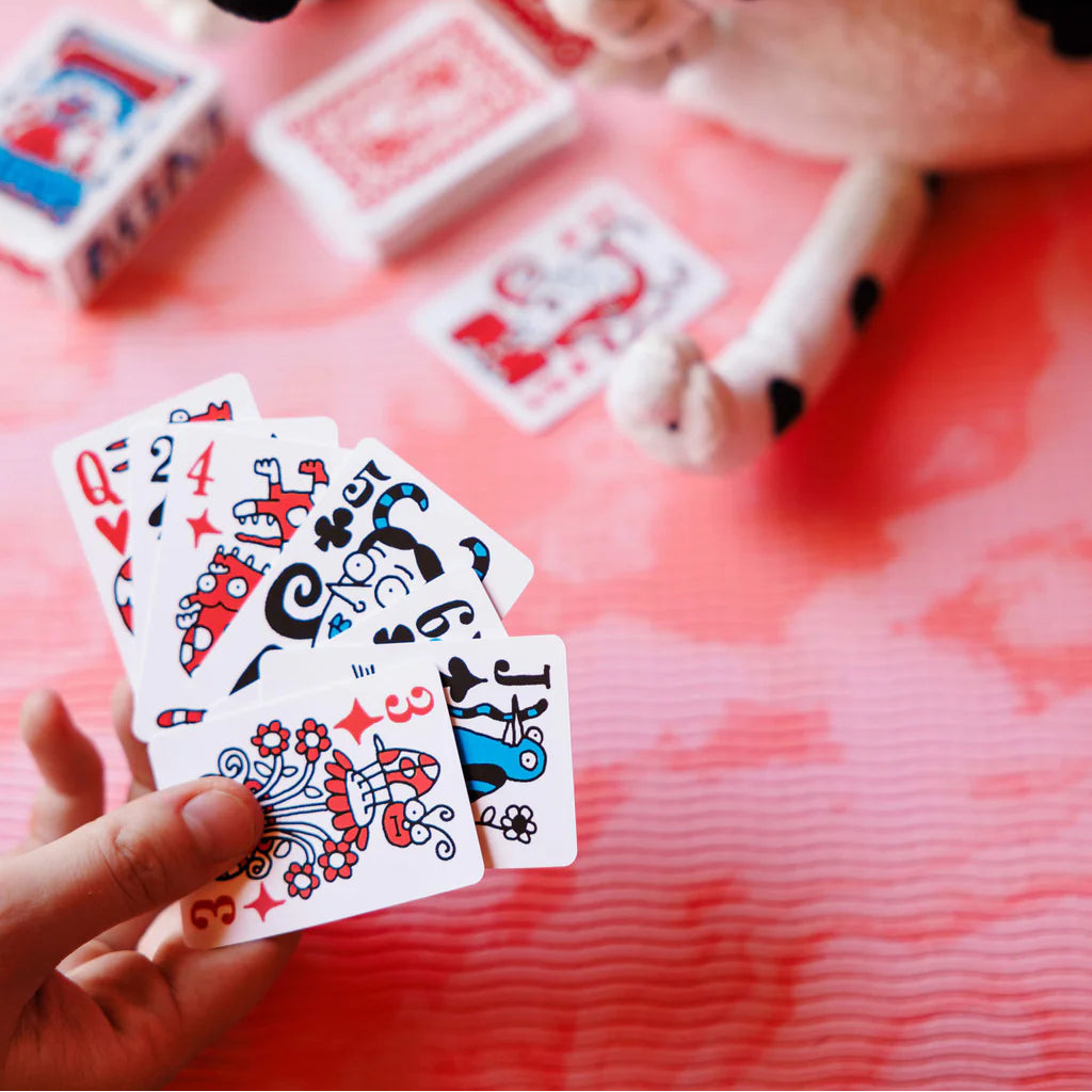 Hand holding playing cards with a cow plush toy and more cards in the background on a pink surface