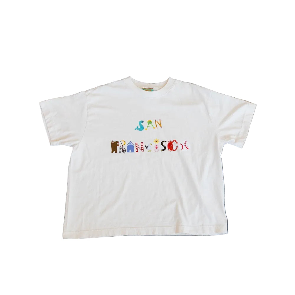 A white short-sleeve t-shirt with a colorful graphic reading 'SAN FRANCISCO' across the chest, laid flat on a textured dark blue and gray concrete surface.
