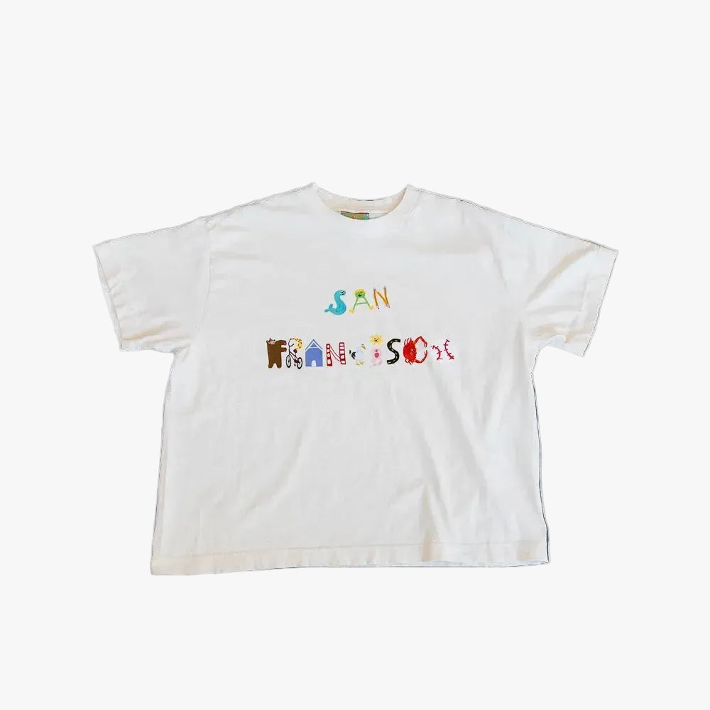 A white short-sleeve t-shirt with a colorful graphic reading 'SAN FRANCISCO' across the chest, laid flat on a textured dark blue and gray concrete surface.