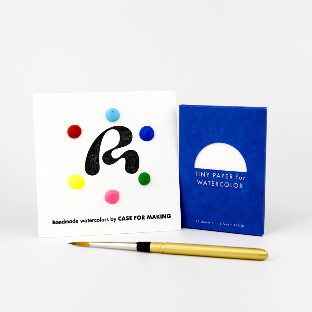 Art supplies including a box of watercolor paper, a brush, and a card with colorful watercolor dots on a white background.