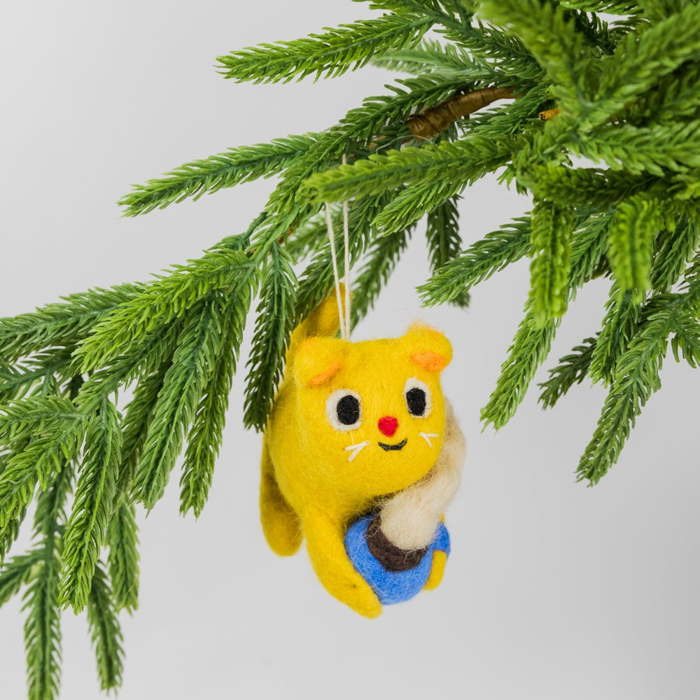 Yellow felted cat ornament holding a steaming cup of cocoa hanging from a tree branch