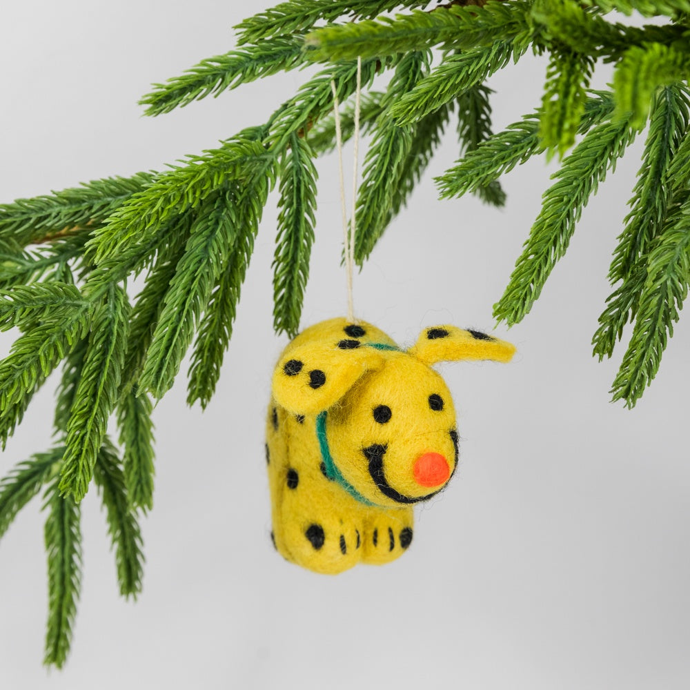 Yellow pooping dog-shaped ornament hanging from a green pine branch on a light gray background