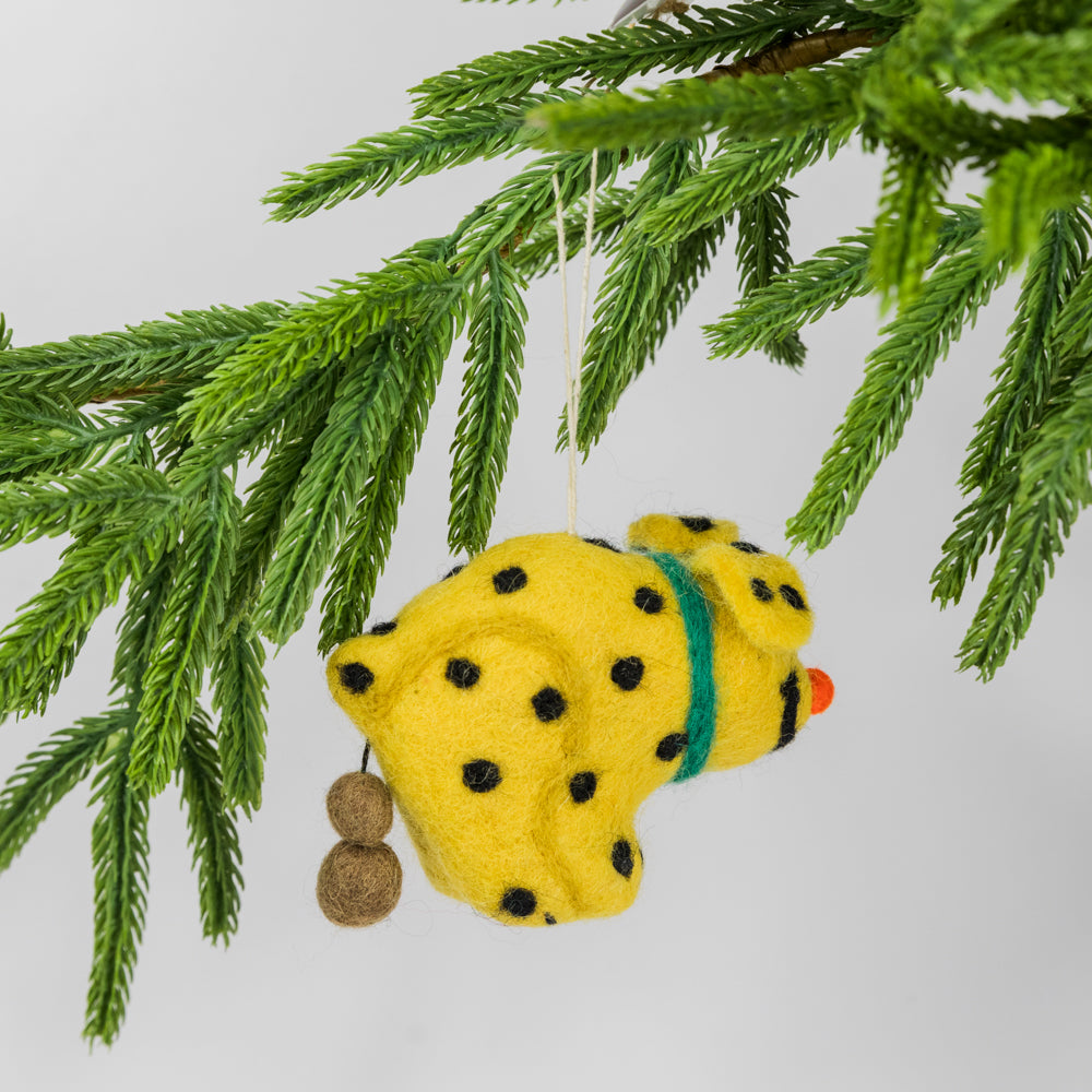 Yellow pooping dog-shaped ornament hanging from a green pine branch on a light gray background
